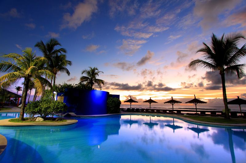 Em um resorts no nordeste, com o céu colorido pelo por-do-sol, uma piscina cercada por coqueiros e guarda sols.
