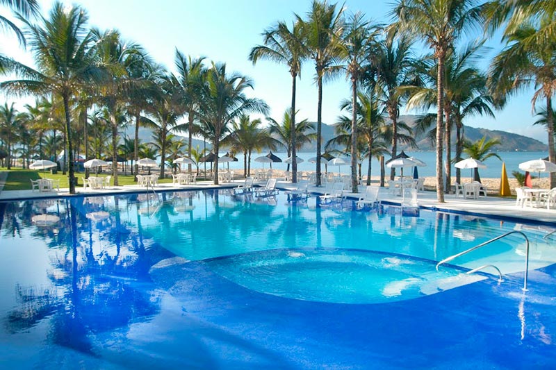 Piscina azulada, cercada por palmeiras e equipamentos de lazer a frente do mar azul em hotel em angra dos reis beira mar