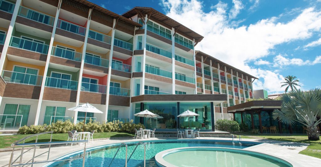 Prédio de poucos andares, pintado de cores claras com uma piscina circular a frente em hotel em porto de galinhas beira mar